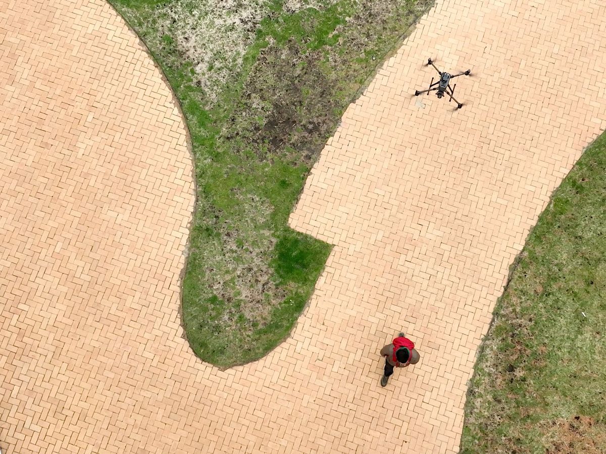 Overhead view of a brick walkway and man with red backpack walking while a drone is flying in the air