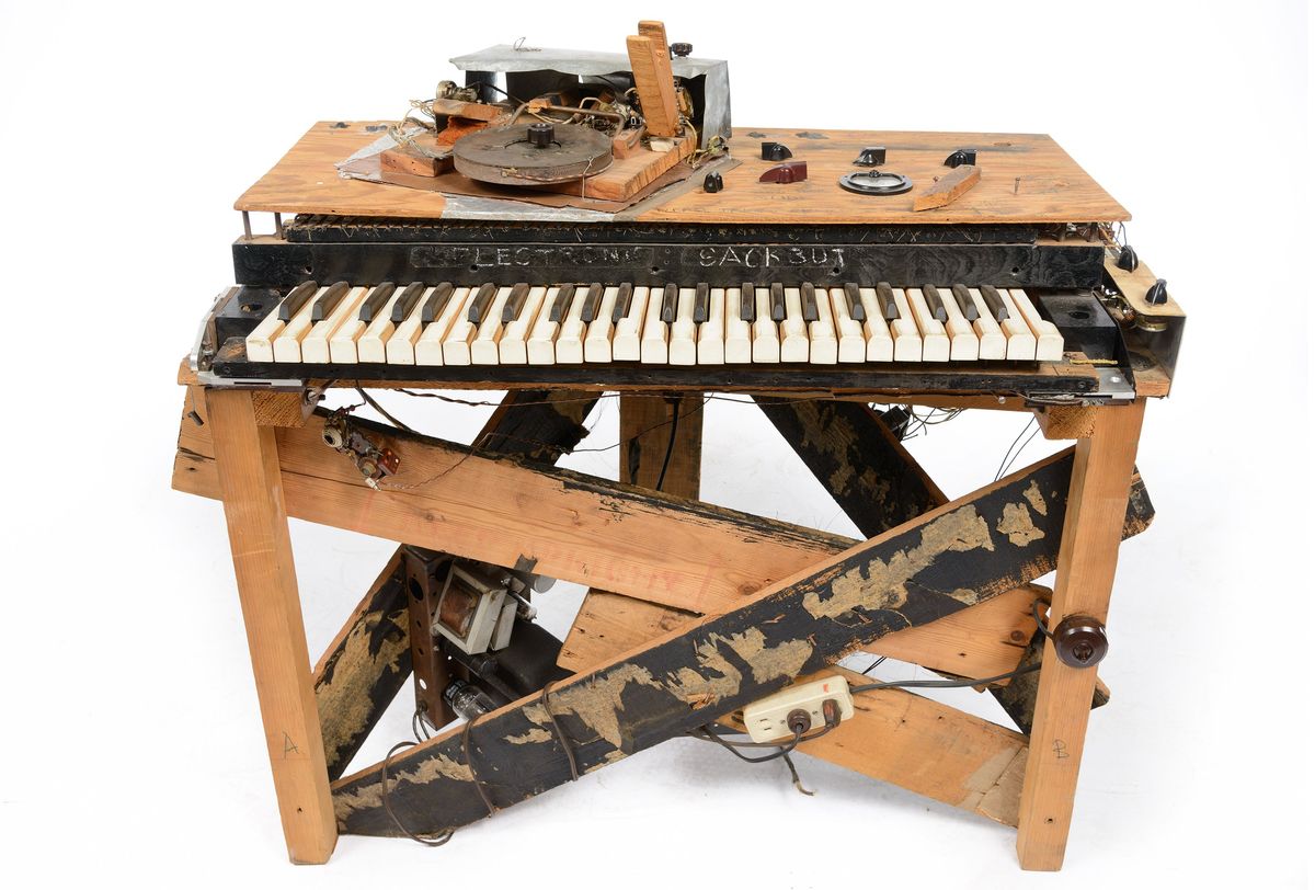 Photo of a crude-looking musical instrument with a keyboard and a rough wooden stand.