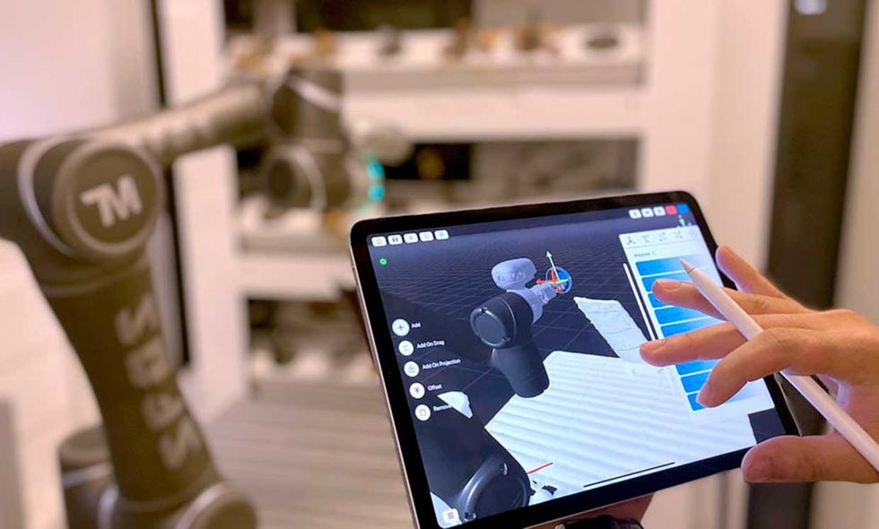 Photo of a hand on a tablet with a robot in the background.  