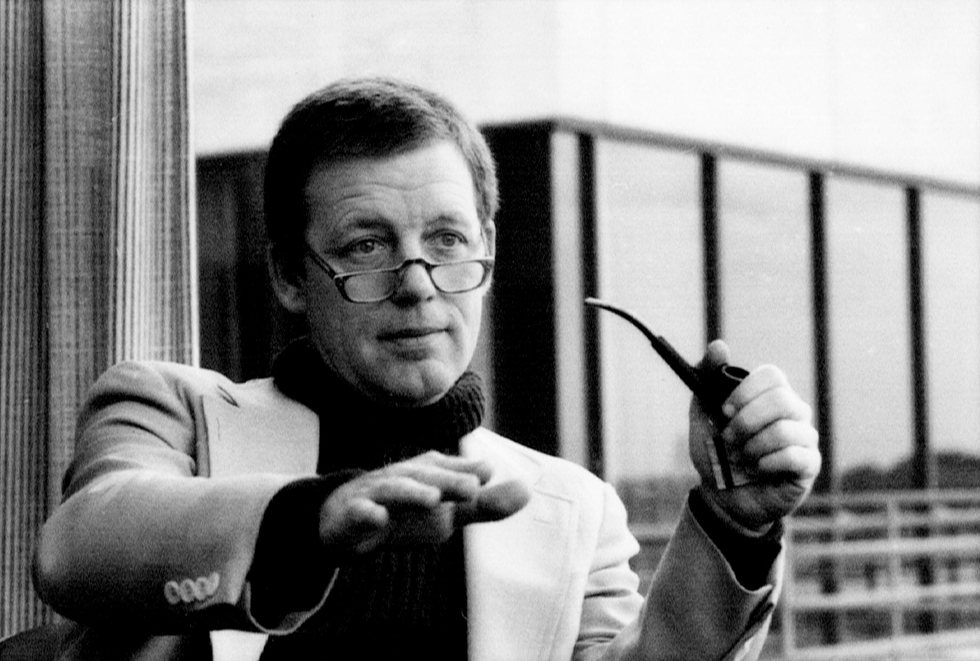 Photo of a man with a turtleneck and glasses, holding a pipe.  