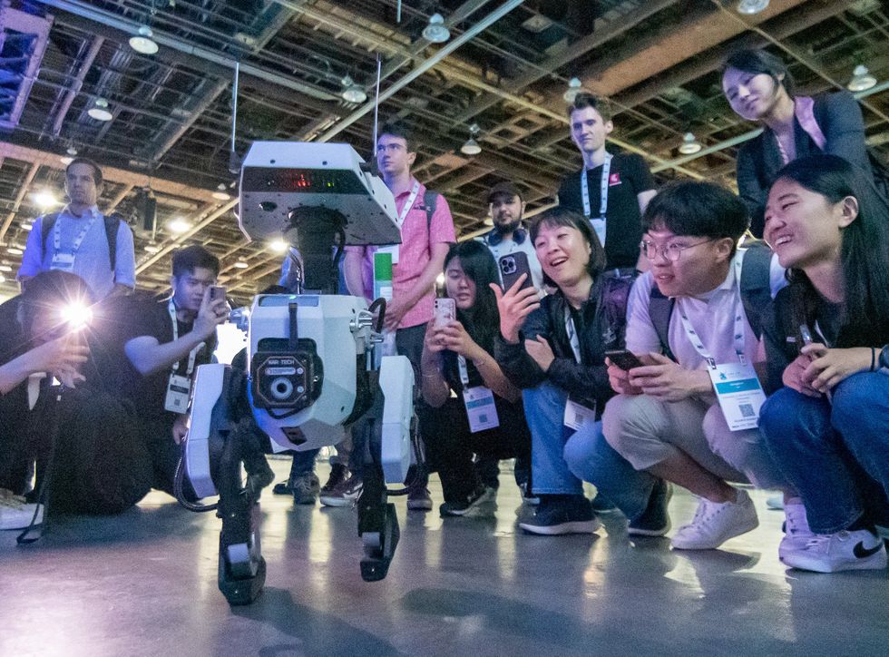 Photo of a small crowd of people surrounding a small robot and taking photos of it.