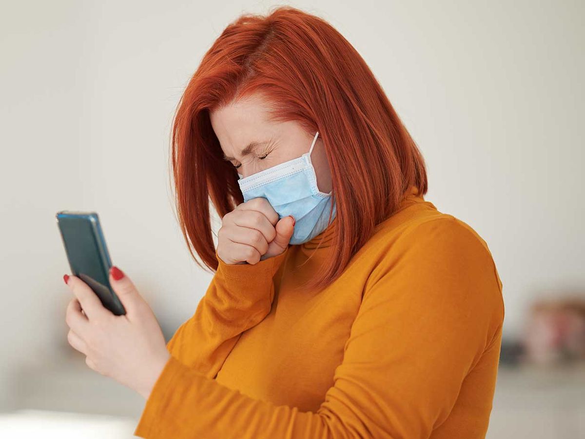 Photo of a woman in a mask, coughing while holding her phone