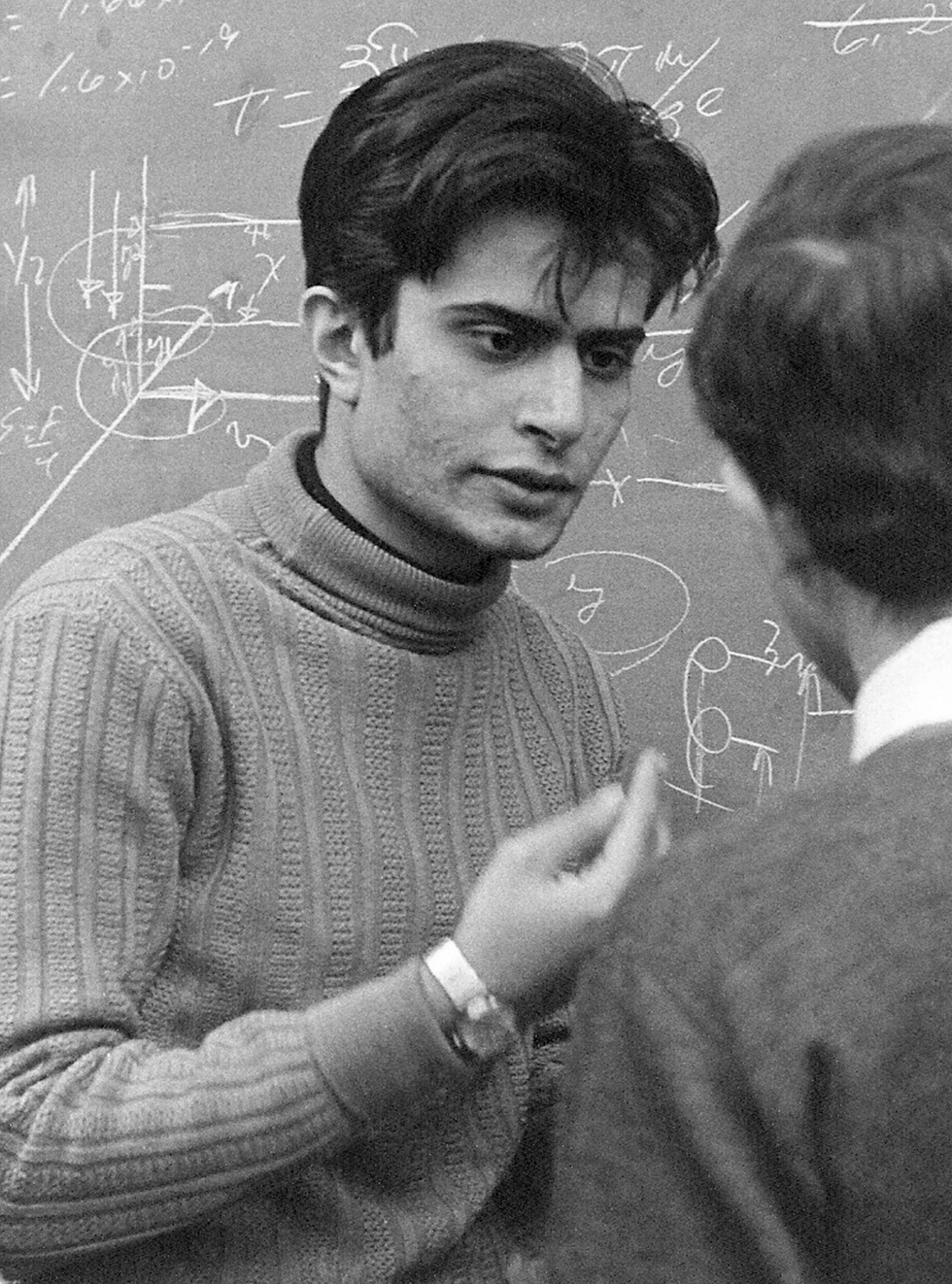 Photo of a young man in a sweater talking to another man in front of a balckboard