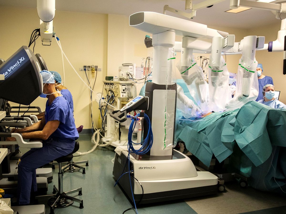 Photo of an operating room. On the left side of the image, two surgeons sit at consoles with their hands on controls. On the right side, a large white robot with four arms operates on a patient.