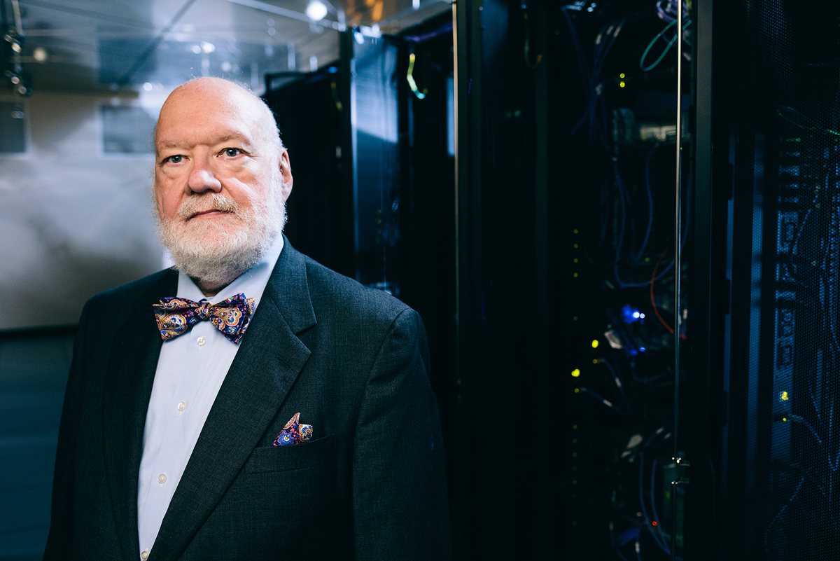 Photo of older white man in a suit standing next to racks of computers.