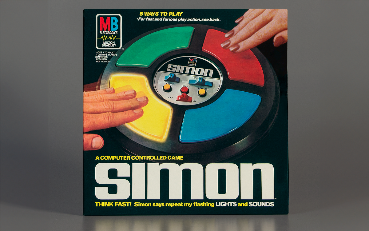 Photo of the box of an electronic game that says “Simon” and shows two hands pressing colorful buttons on a round object. 