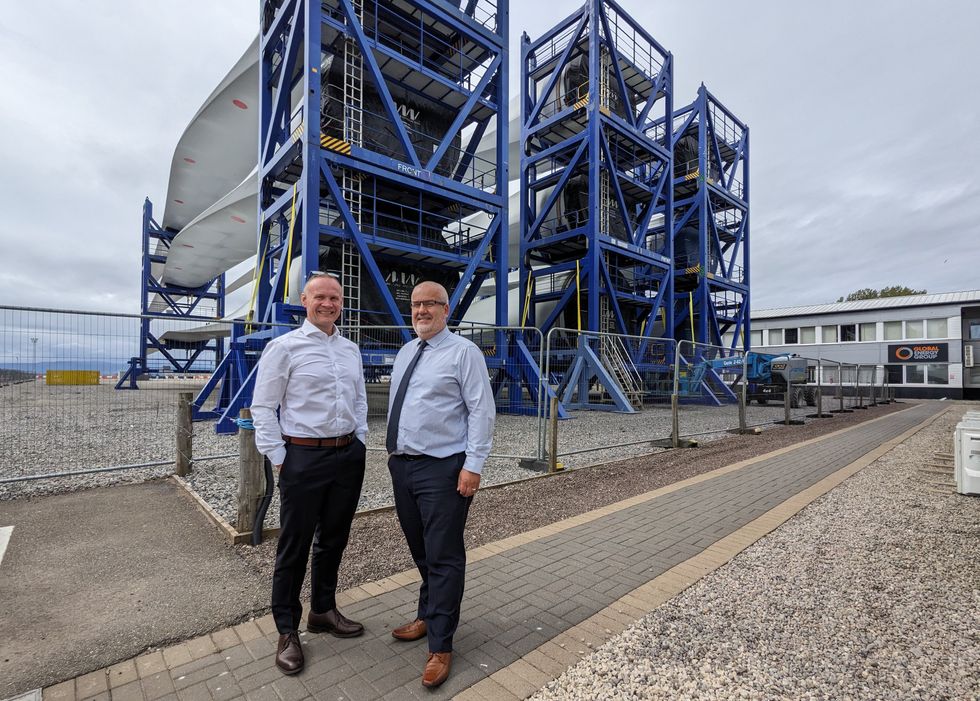Photo of two smiling men standing outdoors in front of giant racks holding the blades of wind turbines. 