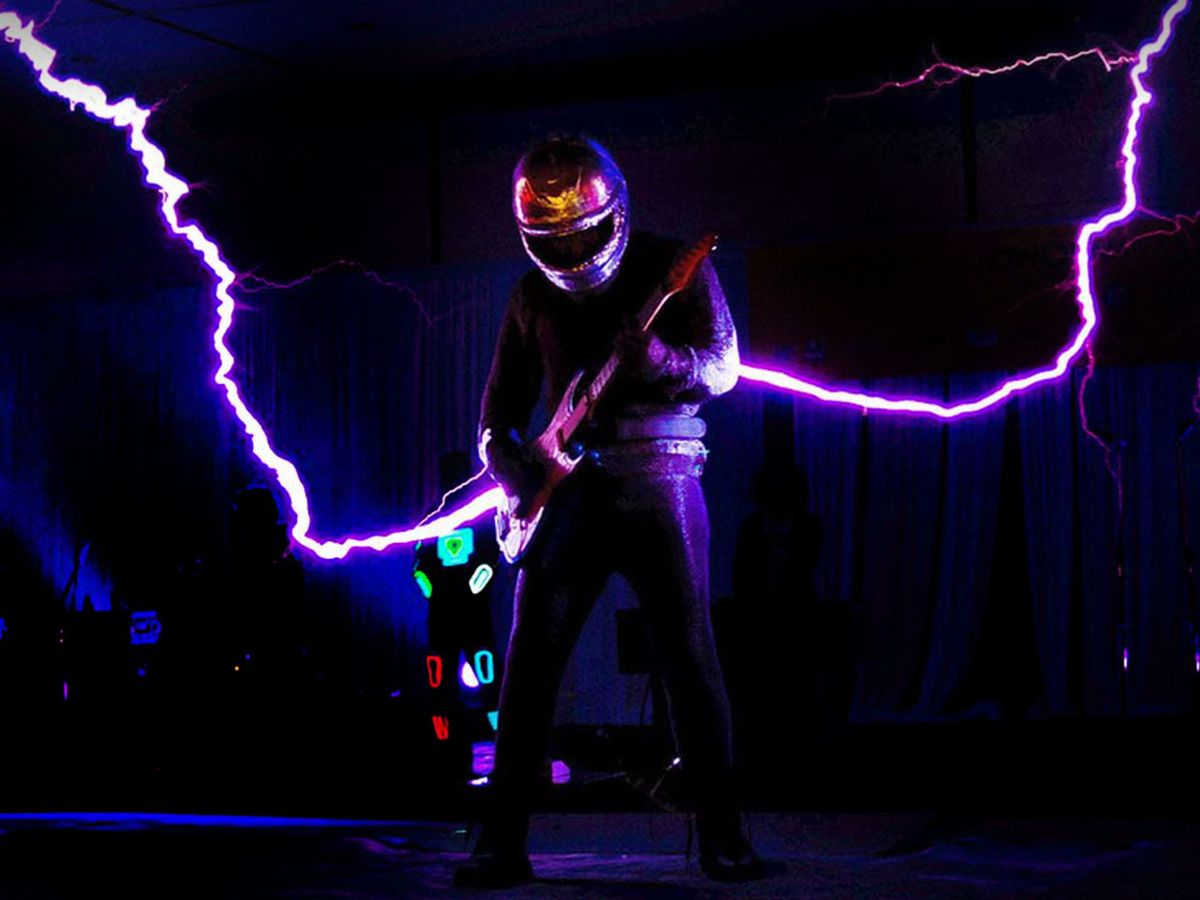 Photograph from an ArcAttack show. A performer is in a wearable Faraday cage and Tesla coils produce giant sparks that look like they are coming out of the guitar.