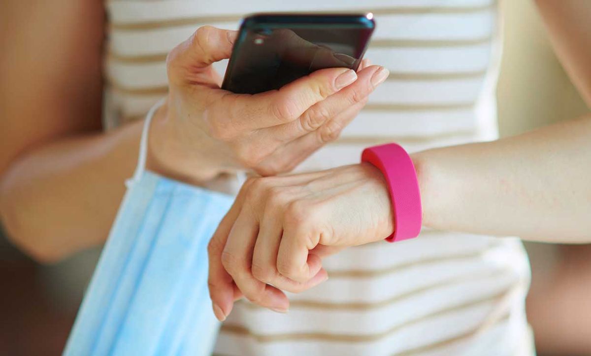 Photograph of a woman's hands. She is holding a mask and a smart phone and has a wearable on her wrist.
