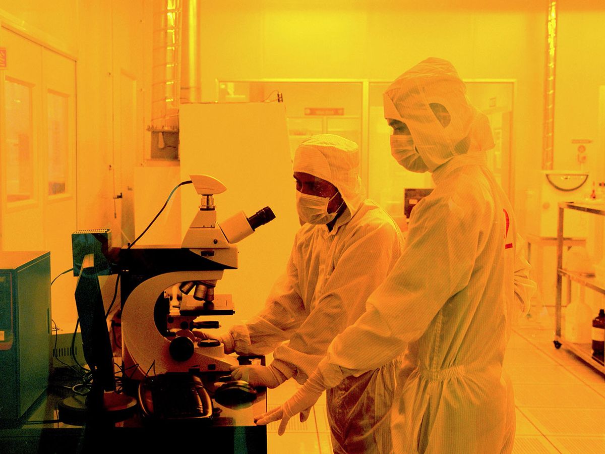 Photograph of two workers wearing bunny suits inside or a semiconductor fabrication lab.