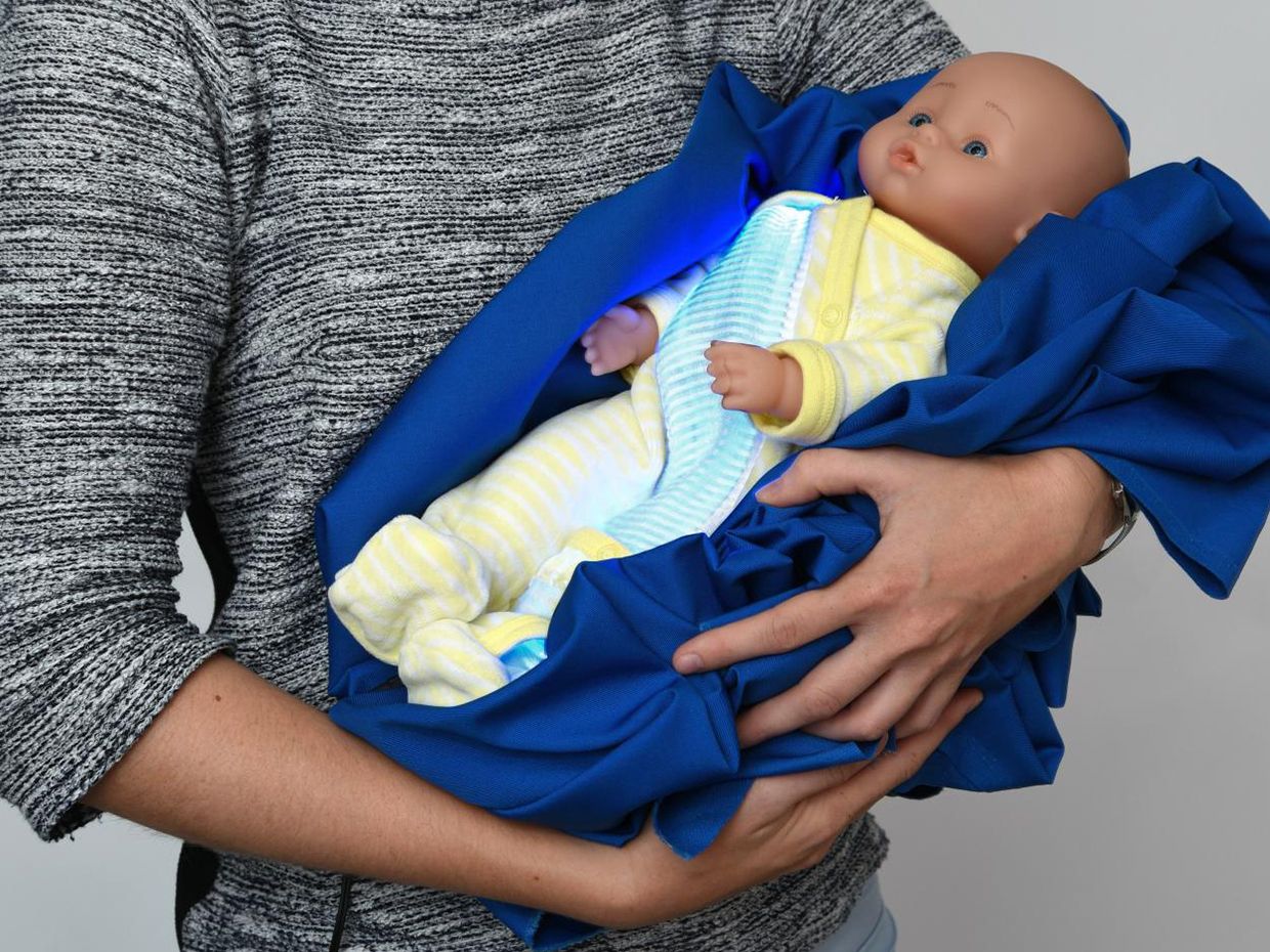Photograph showing a woman holding a baby doll in a blue blanket an a yellow onesie, with a strip of blue-light illuminated textile along the front.