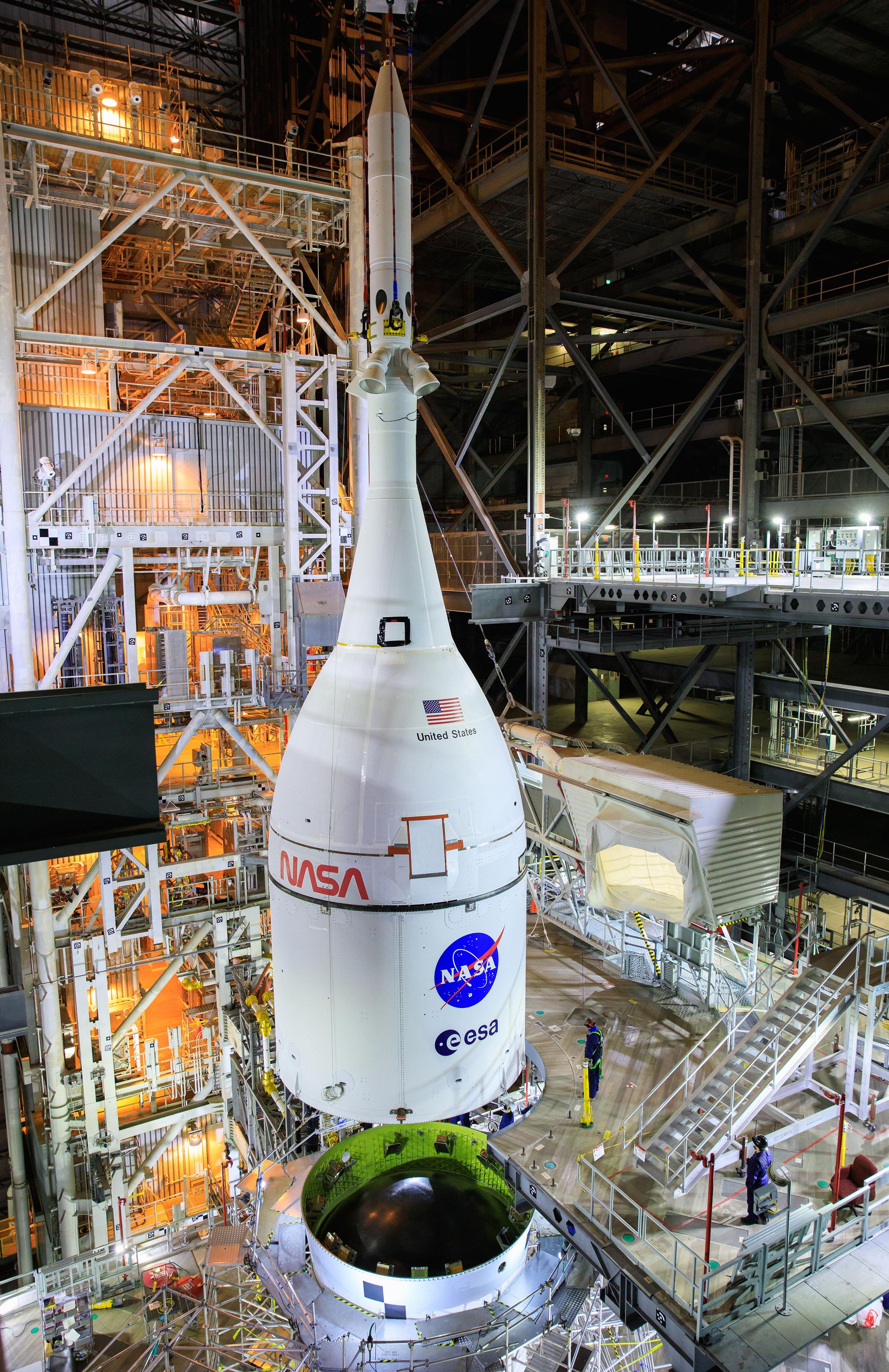 Photograph shows a bullet-shaped spacecraft suspended just above the top of the Space Launch System booster, inside a cavernous building.