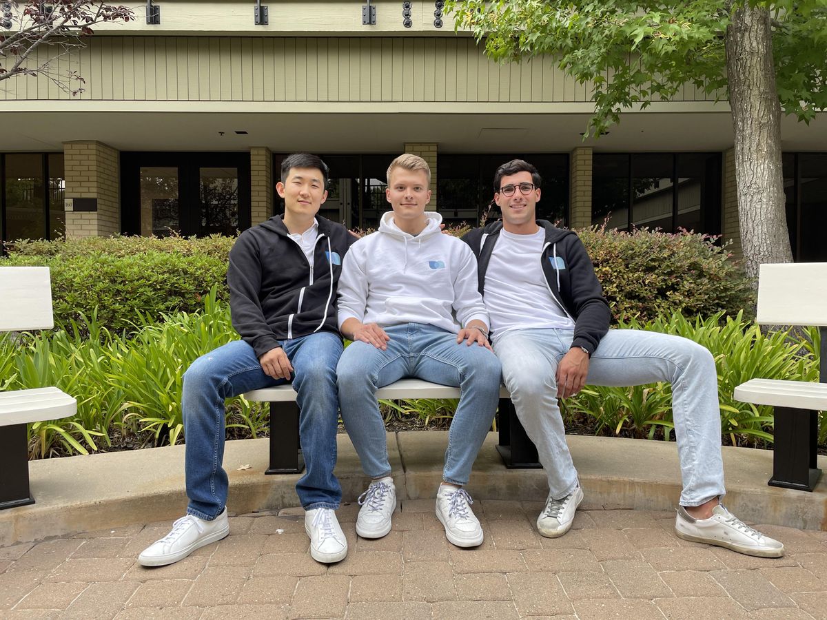 Picture of three young men sitting outside on a bench in front of bushes and greenery