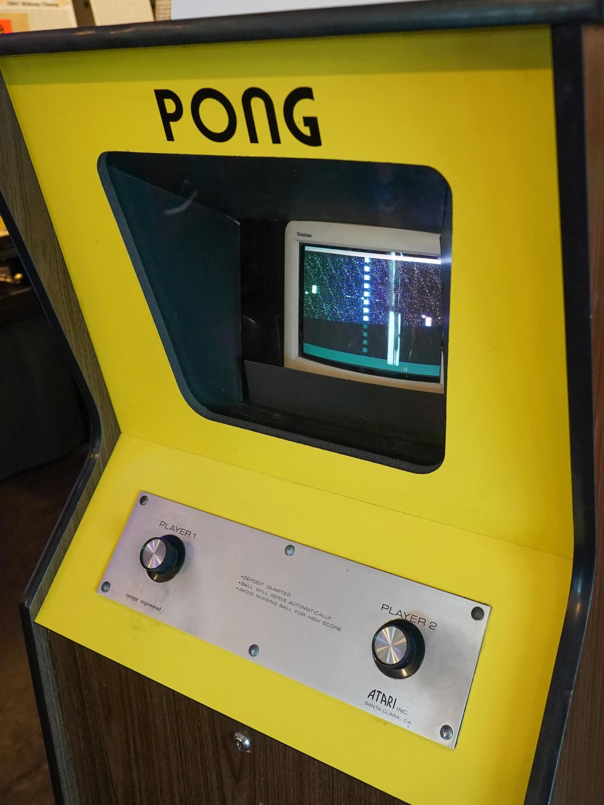 Pong arcade game in yellow cabinet containing black and white TV display, two knobs are labeled Player 1 and Player 2, Atari logo visible.
