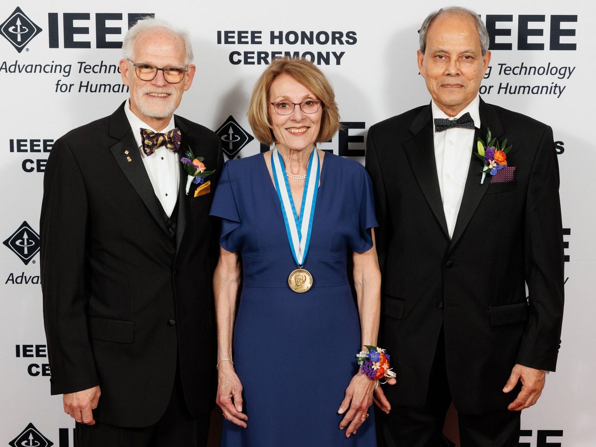 portrait of 2 men in black suits and a woman in a navy blue dress and a medal around her neck smiling at the camera.
