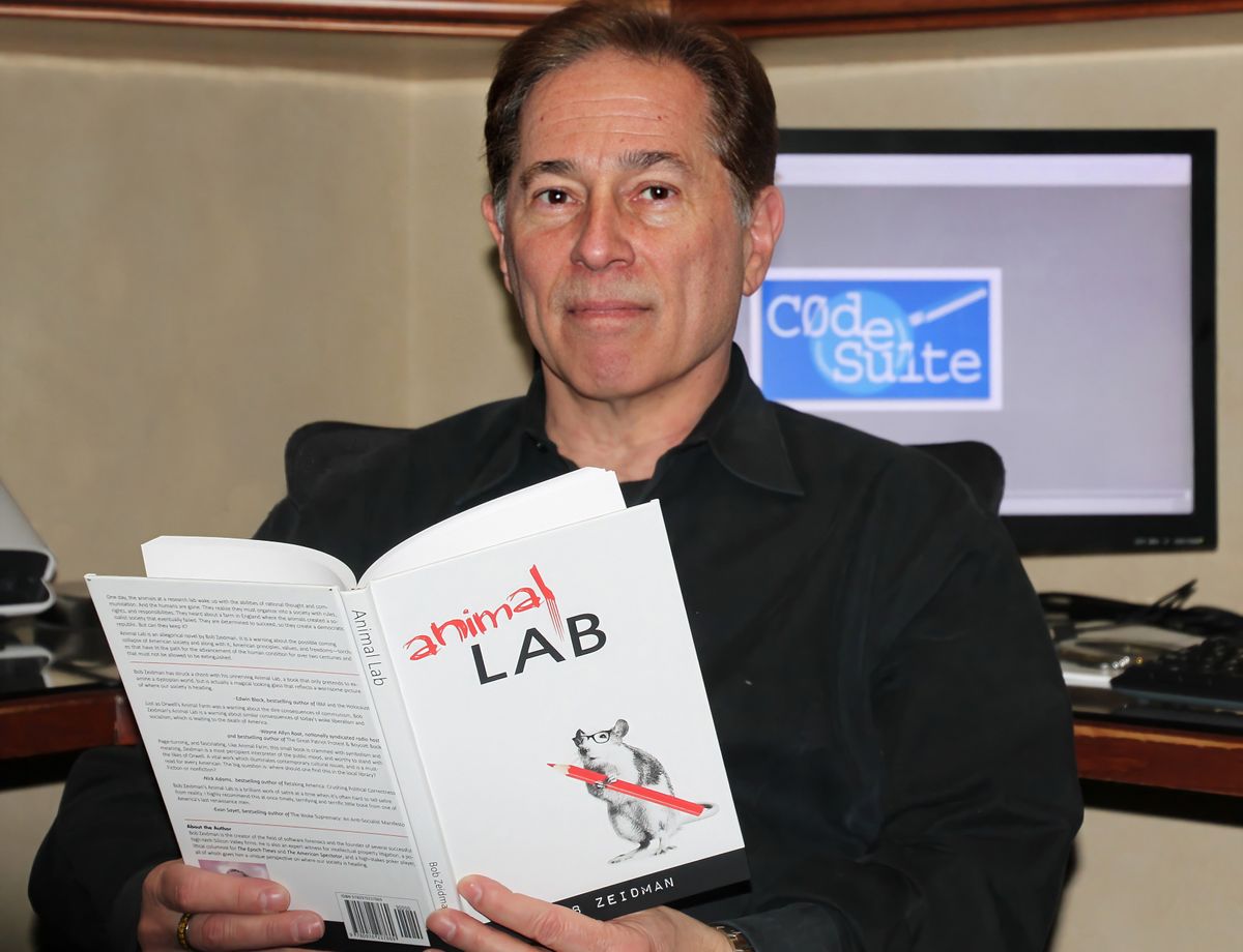 Portrait of a man in a black shirt sits at a desk holding a book called Animal Lab. The computer behind him says Code Suite.