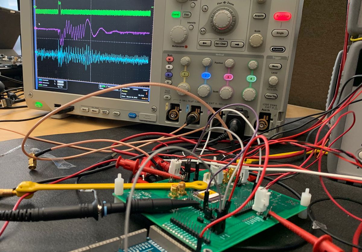 Probe wires are attached to a green circuit board. An oscilloscope displays multiple traces in the background.