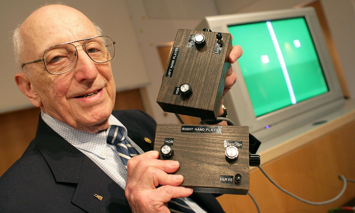 Ralph Baer, a smiling older man in glasses, holds two brown boxes with nobs. In the background a screen displays white shapes on a green background.