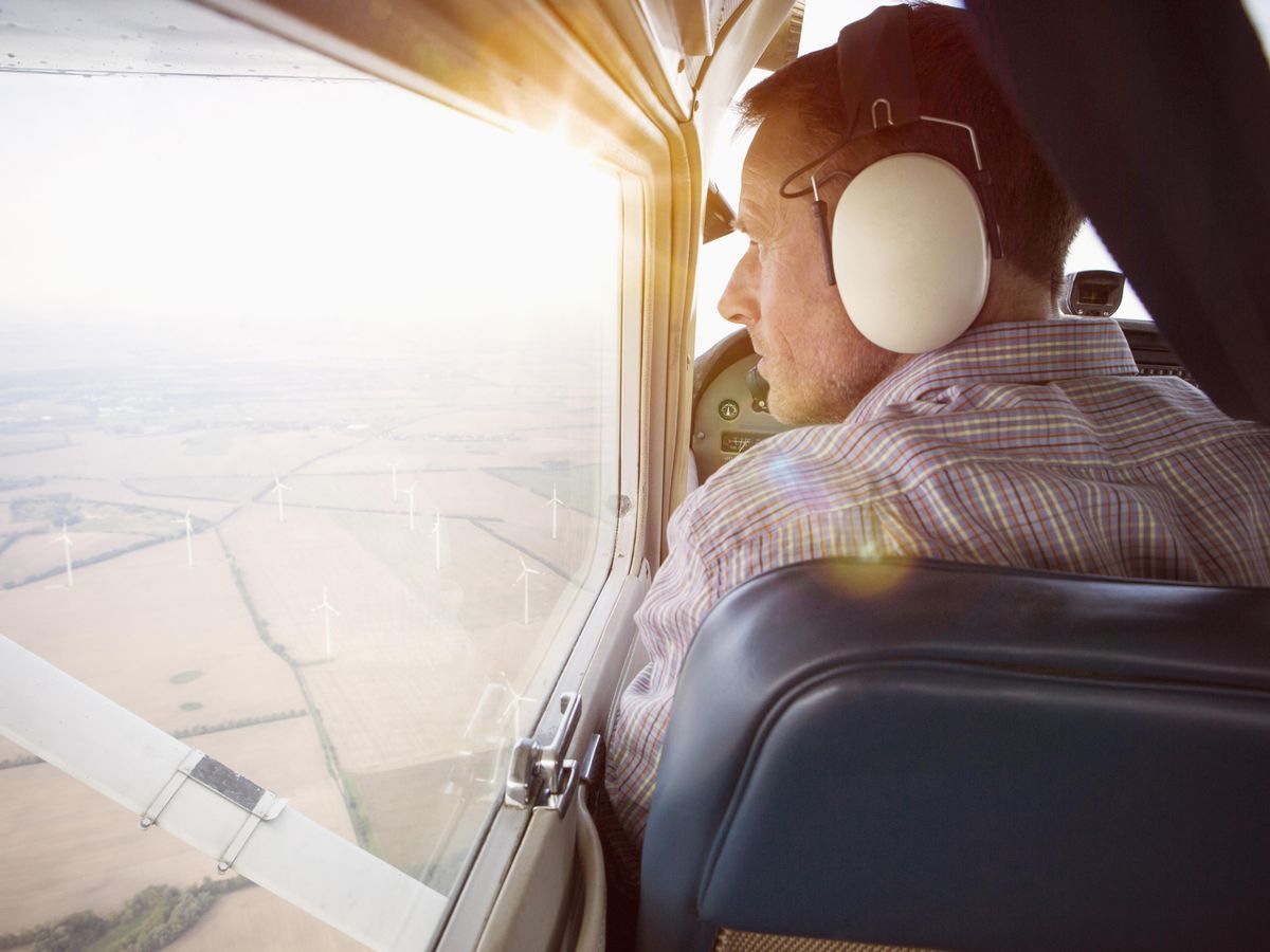 Rear view of a man with headphones looking out of the window from a private plane. Below is an aerial view of land
