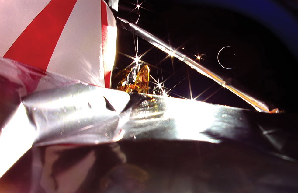Red, white and silver parts of a spacecraft in the foreground, with a delicate crescent Earth beyond, framed against black sky.