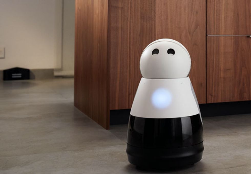 Robot with round white head and a coned shaped body sits on the floor in front of wooden kitchen cabinets