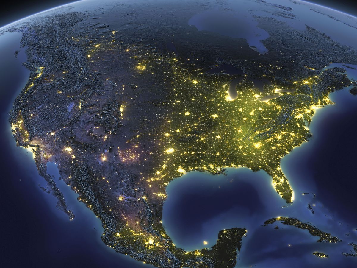 Satellite overhead view of the United States at night. The land mass is grey with bright yellow lights across the country. 