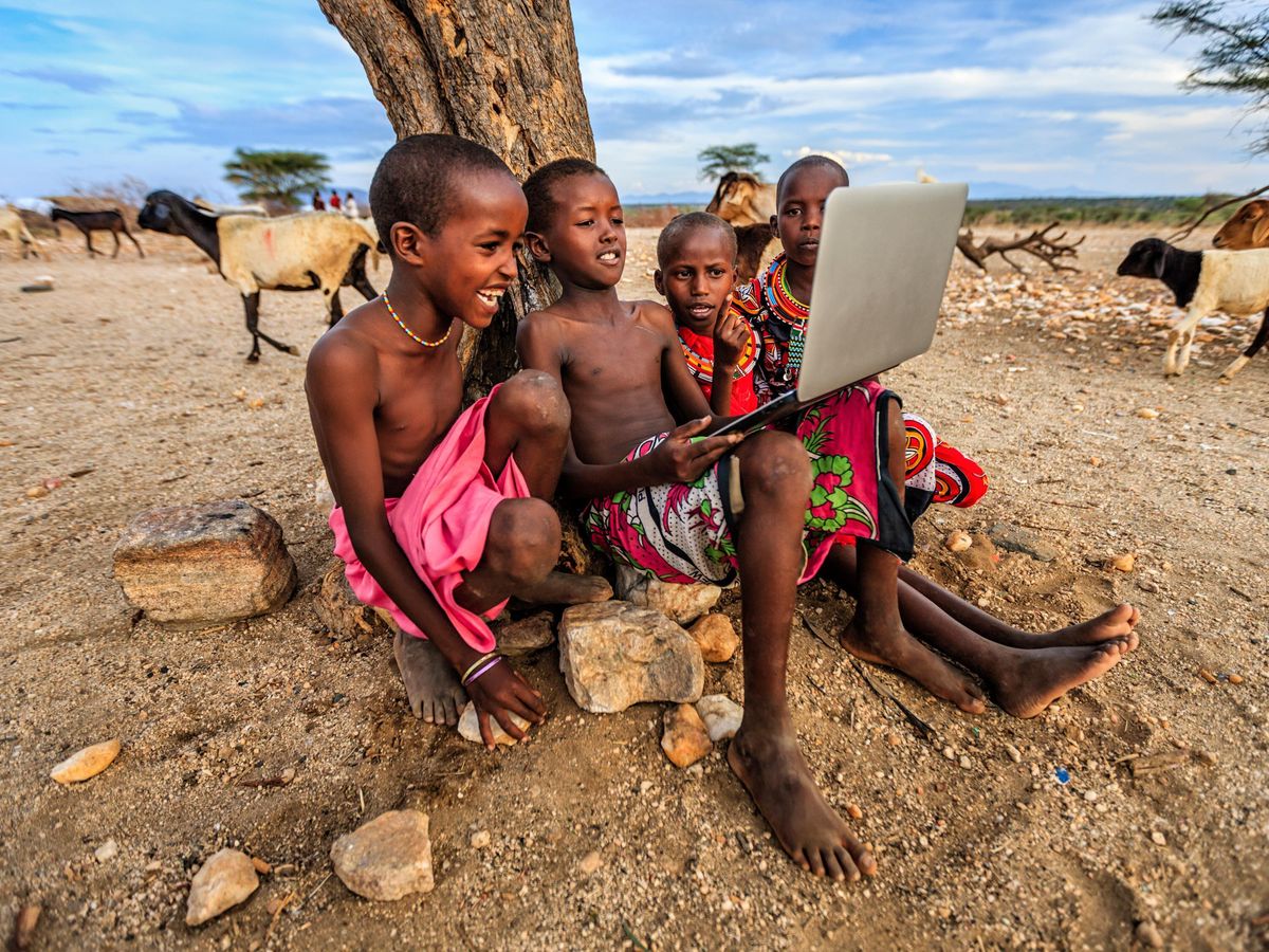 Several children in traditional clothing sitting by a tree looking at a laptop.  
