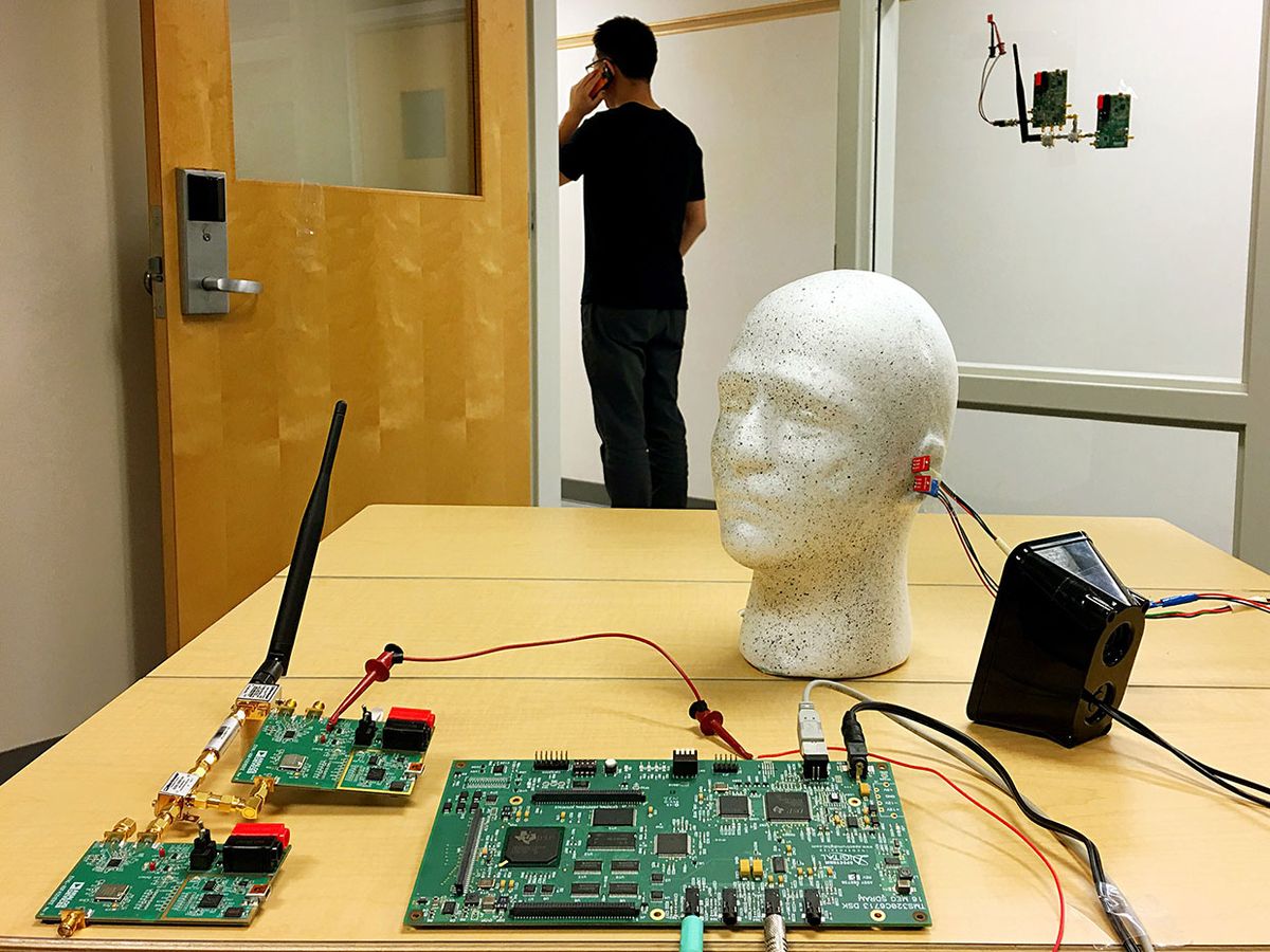 Several circuit boards on an office desk are connected with wires to a loudspeaker. A dummy head has sensors attached to its ears.