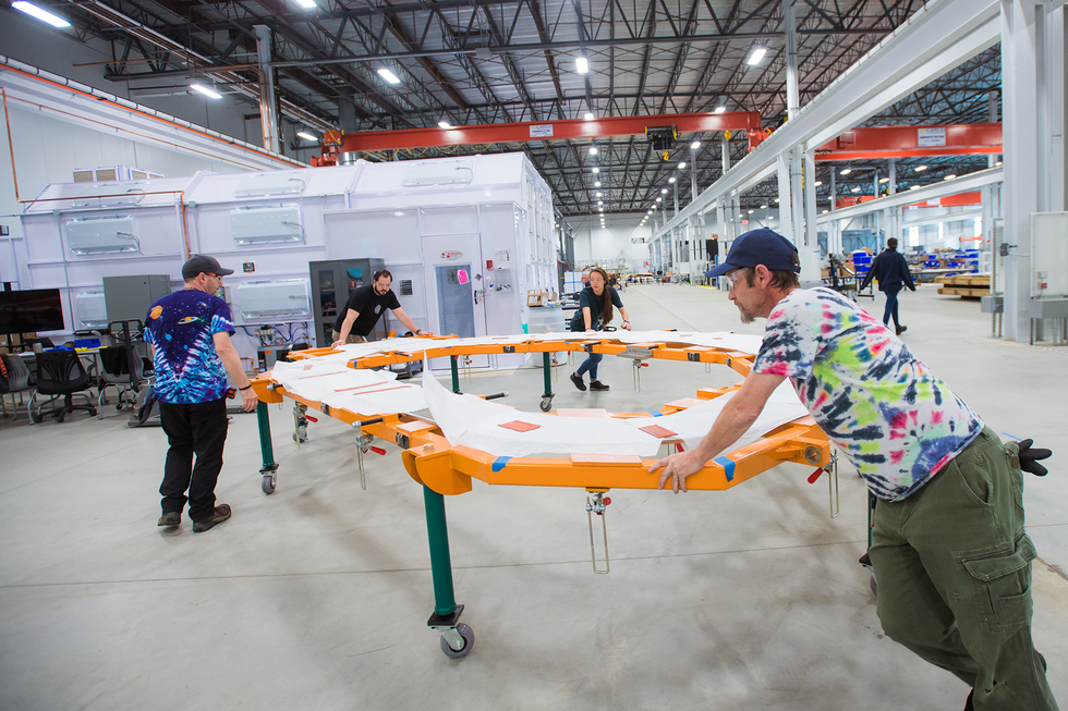 Several people navigate a D-shaped white and orange surface on wheels through a large industrial space.