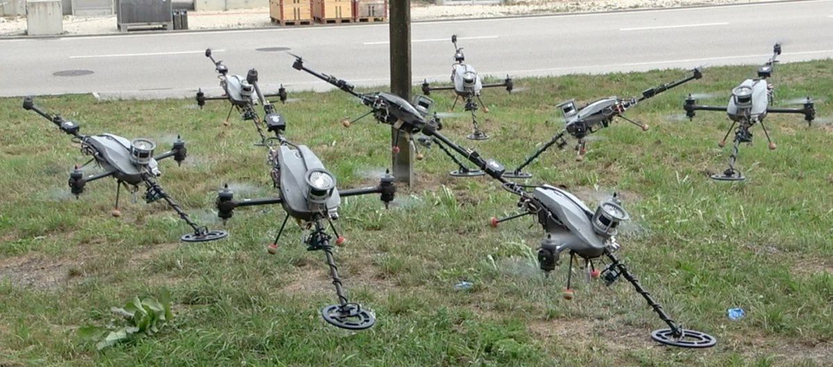 Several stitched-together photographs of gray three-propellered drones with metal detectors hovering just above grass