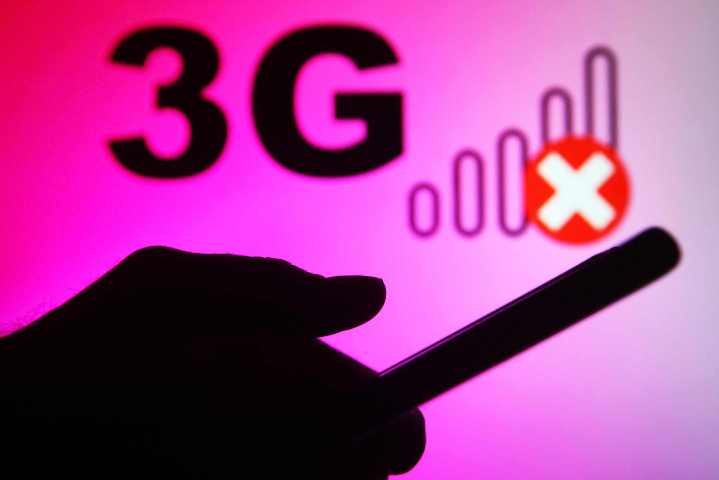 Silhouette of a hand holding a phone in front of a pink backdrop that says 3G and has empty bars to indicate no signal. 
