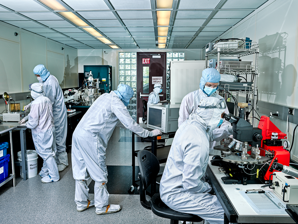Six people , three in foreground and three in background, work with laboratory equipment. They are all dressed in white jumpsuits and wear head coverings and facemasks.  