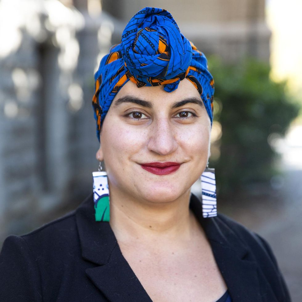 Smiling woman in a blue and orange head scarf and large rectangular earrings.