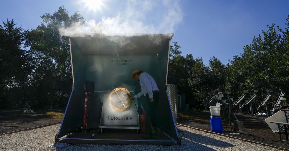 Steam rises from a giant coffee roaster that is being powered by the sun. For scale, one of its inventors stands in front of it; he\u2019s just over half the machine\u2019s height.