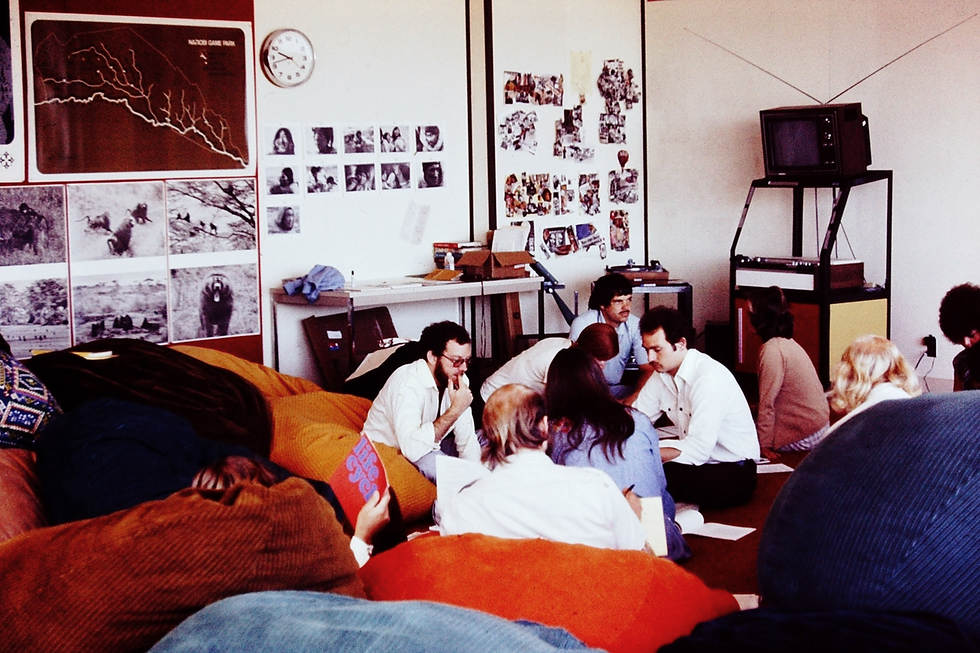 Ten people, most dressed in button-down shirts without ties, sit on red, blue, and orange beanbags scattered on the floor of a room. Much of the wall space is covered with bulletin boards and white boards scattered with photographs. 