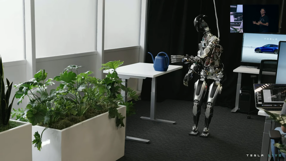 The humanoid robot stands near a desk and extends its arm to grab a watering can.