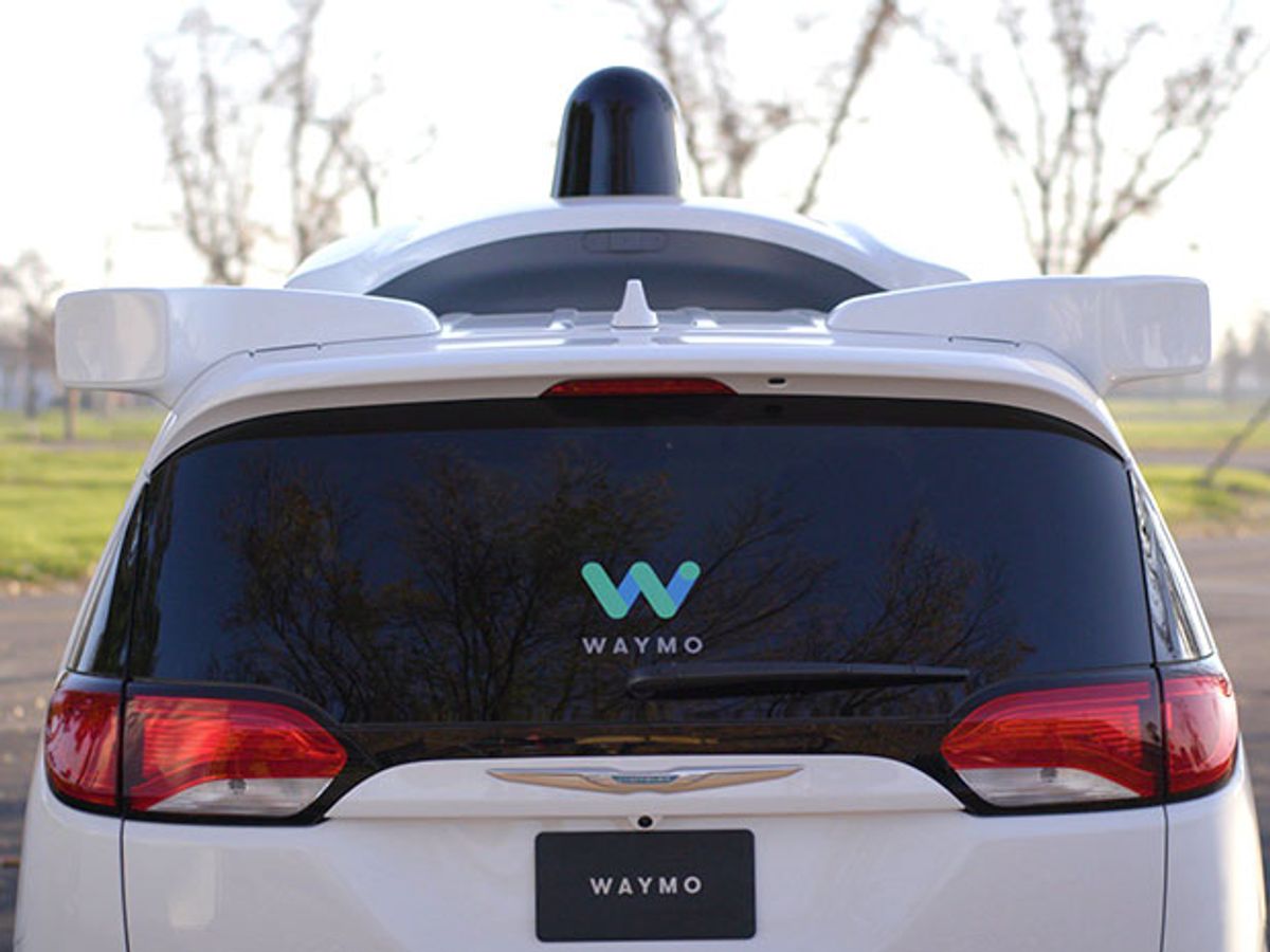 The rear end of a white car with a black knob on top and sign that says “Waymo” on the rear windshield