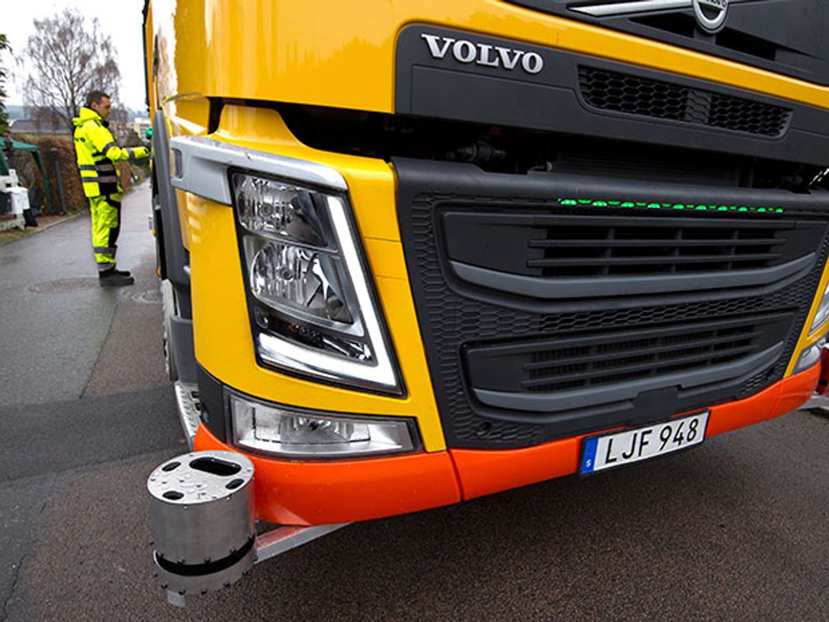 The right front corner of a yellow garbage truck. A metallic lidar cylinder sticks out at the corner. A man in neon yellow stands in the background.