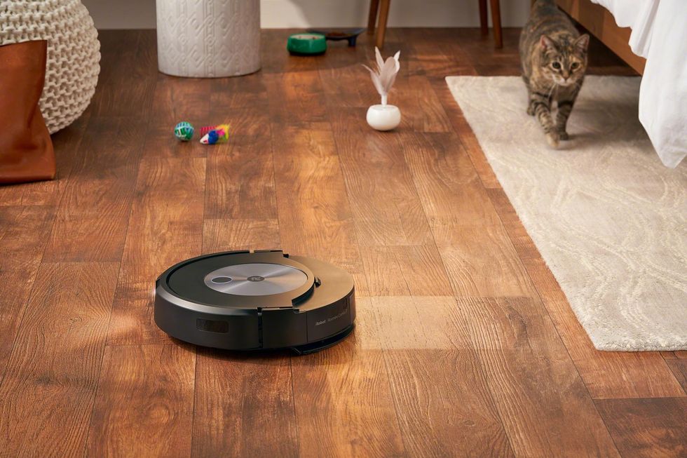The robot mopping a hardwood floor with scattered cat toys around it and a cat in the background