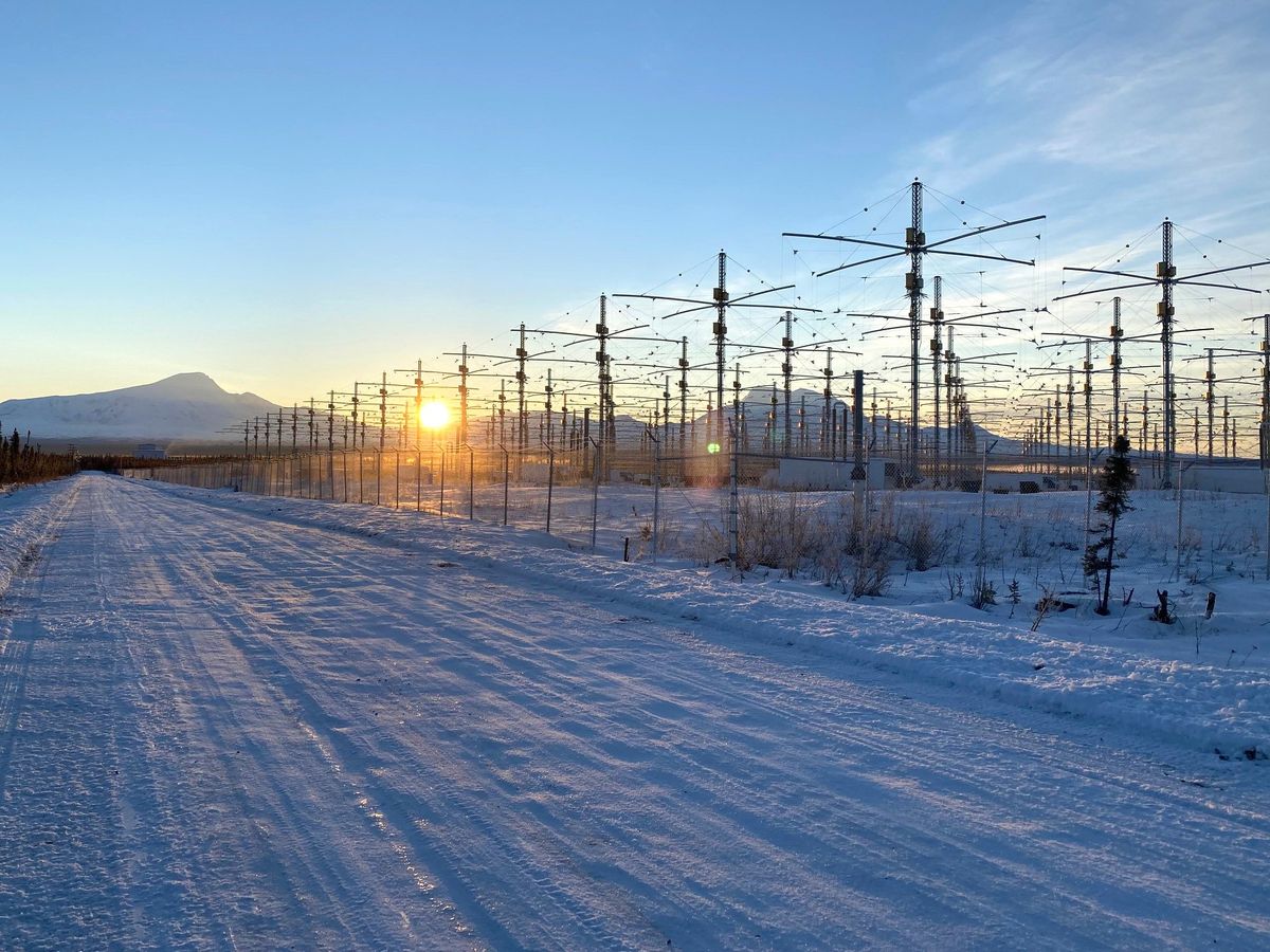 The sun peeks over a distant mountain range, while in the foreground lies a snow-covered road and a field of identical antennas, each a vertical pole topped by two crossbars.