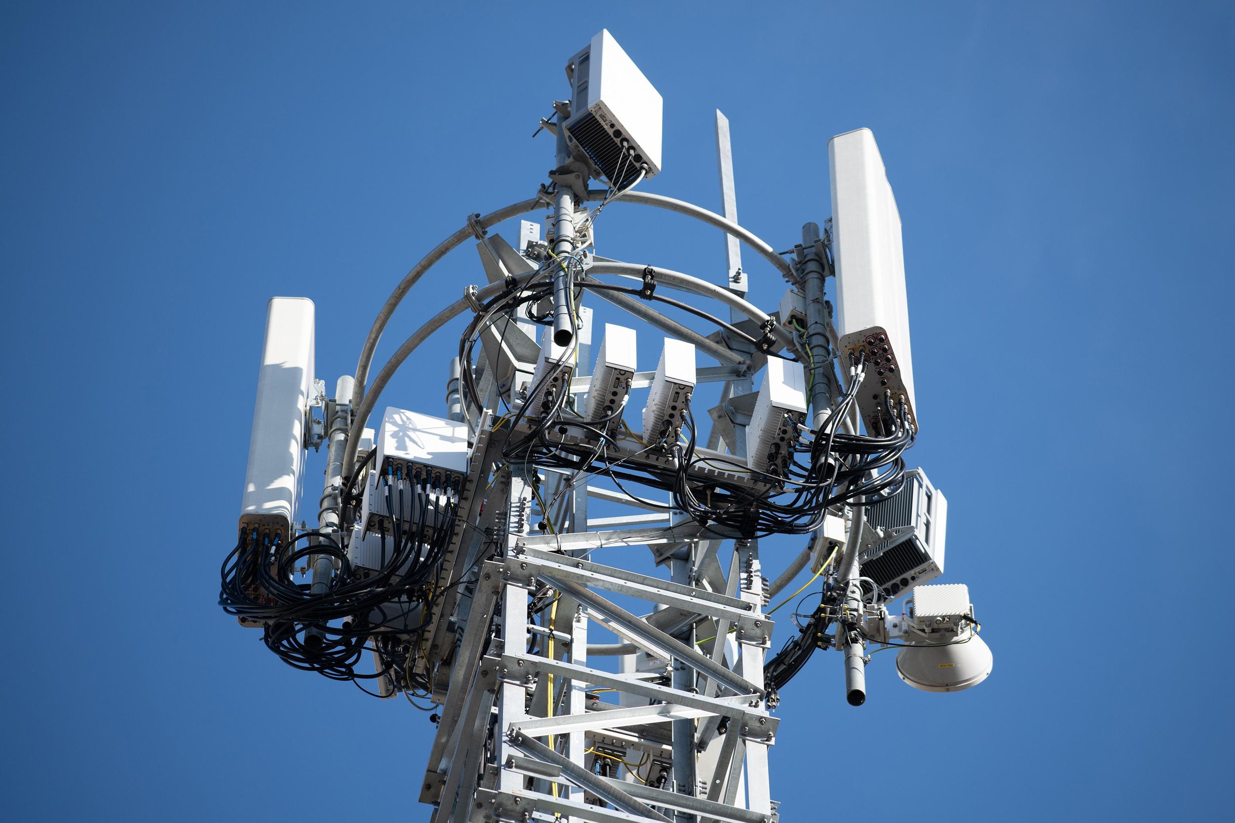 The top of a mobile phone mast covered in telecommunications equipment is seen against a clear blue sky.