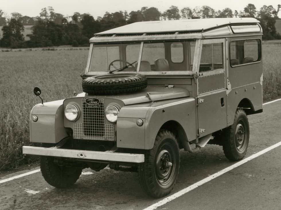 This black-and-white image shows a 1954 Land Rover Series 1 model, a boxy vehicle with a flat windshield and its spare tire mounted on the hood. 