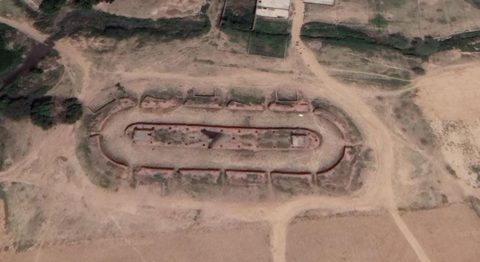 This image shows a race-track shaped structure with a tall chimney in the middle, built in an area where the ground is a distinctly reddish hue.