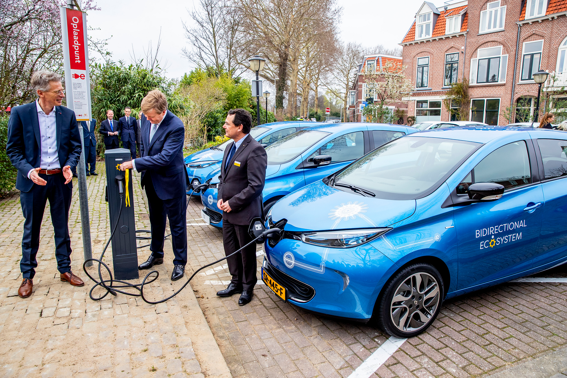 This image shows three men in suits standing next to a charging station that is charging a blue electric car with the words \u201cBidirectional Ecosystem\u201d written on the door.