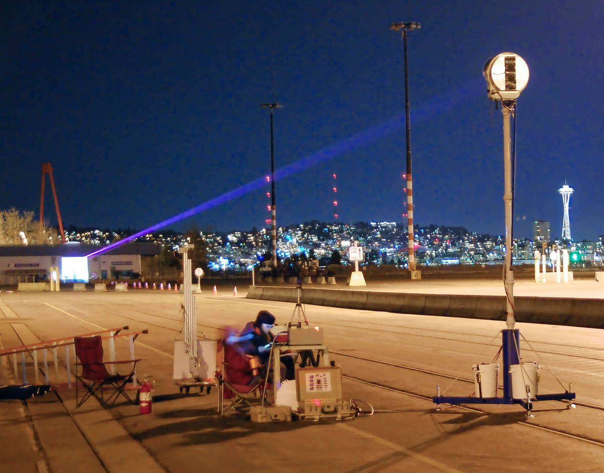 This nighttime outdoor image, with city lights in the background, shows a narrow beam of light shining on a circular receiver that is positioned on the top of a pole.