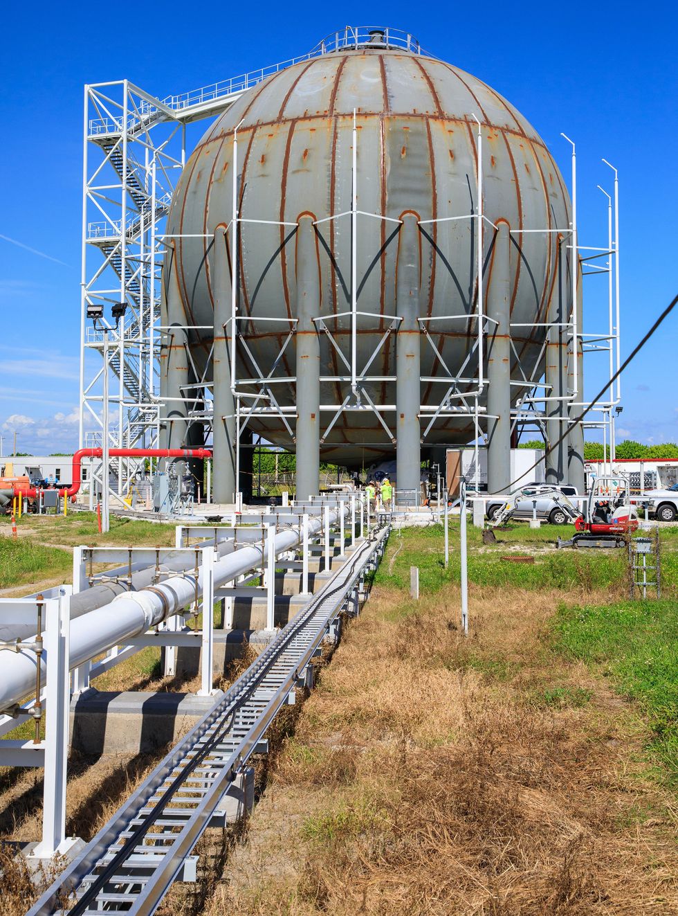This photograph shows a giant spherical storage tank with an adjacent stairway to the top and pipes leading to it that are close to the ground.
