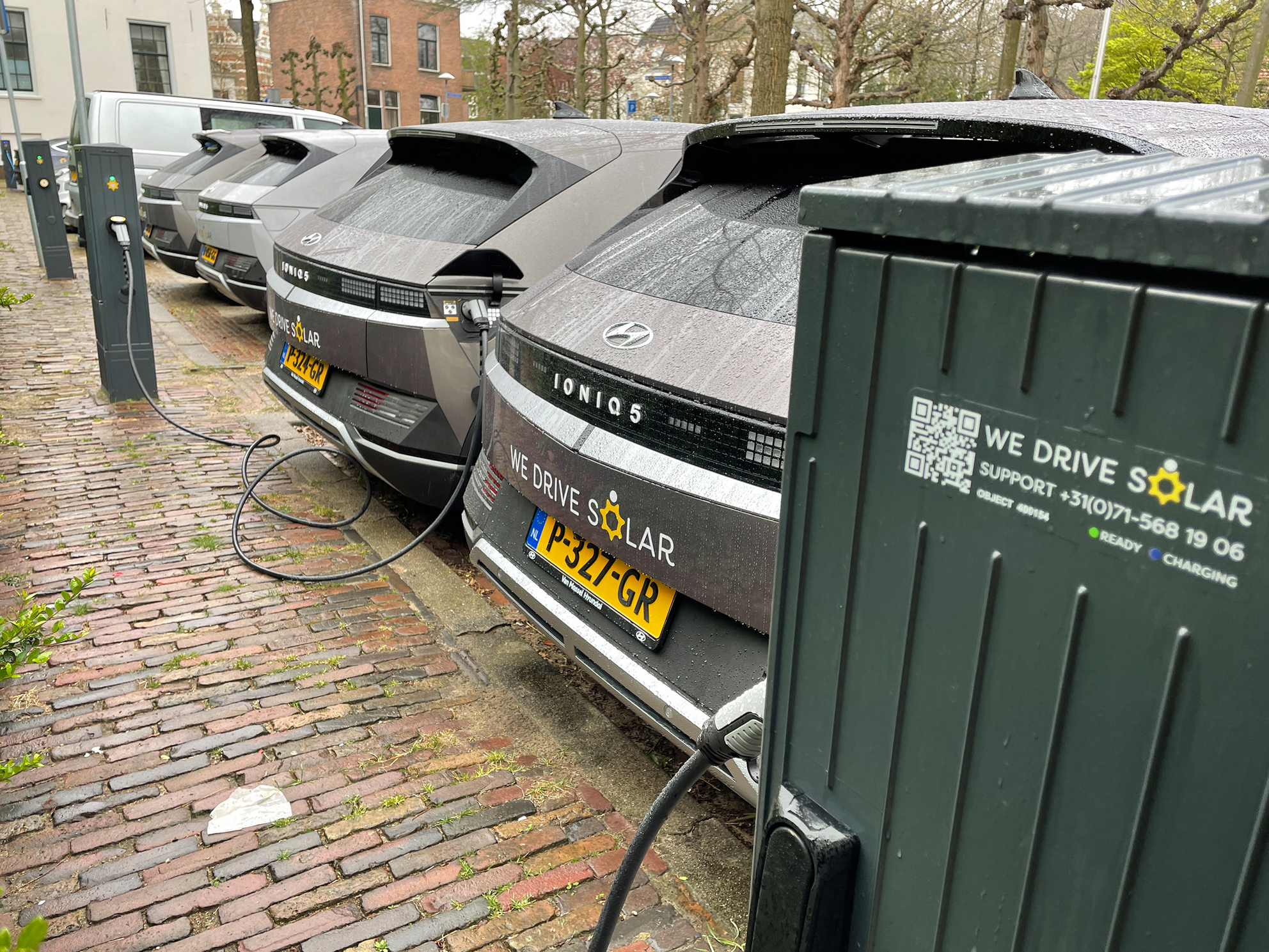 This photograph shows four parked vehicles, each with the words \u201cWe Drive Solar\u201d prominently displayed, and each plugged into a charge point.