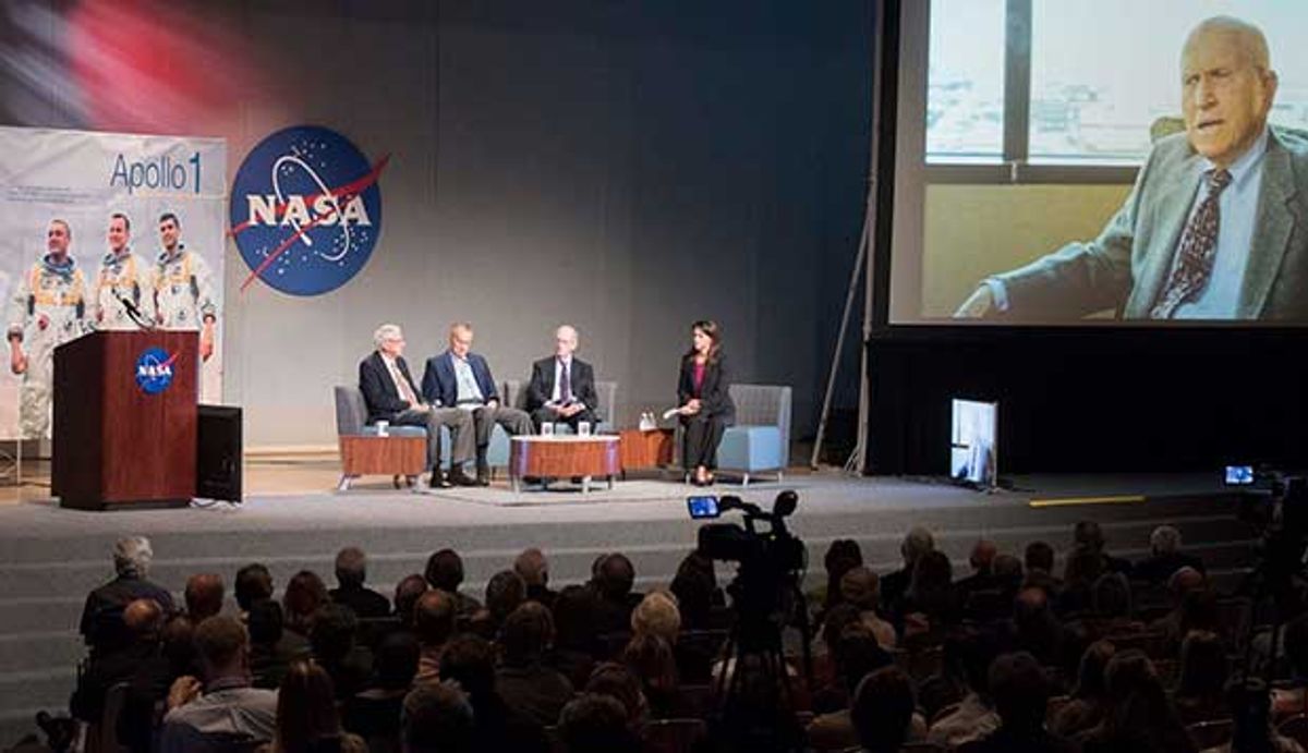 Three elderly male panelists and one younger female moderator sit on a stage above an audience. Another eldery man can be seen on a large video screen.