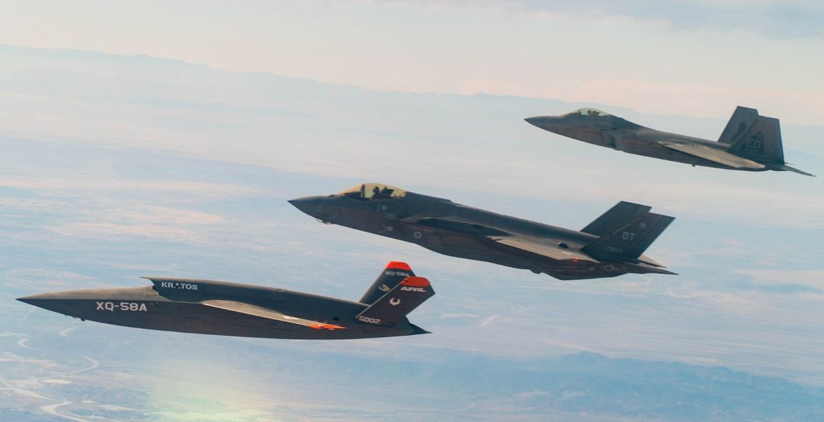Three fighter jets fly in the air. The one in the foreground does not have a cockpit like the other two, and is labelled XQ-58A.