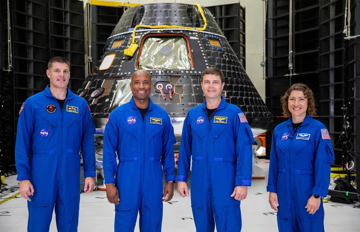 Three men and a woman, all dressed in blue uniforms, stand abreast and in front of a large, semiconical space capsule.